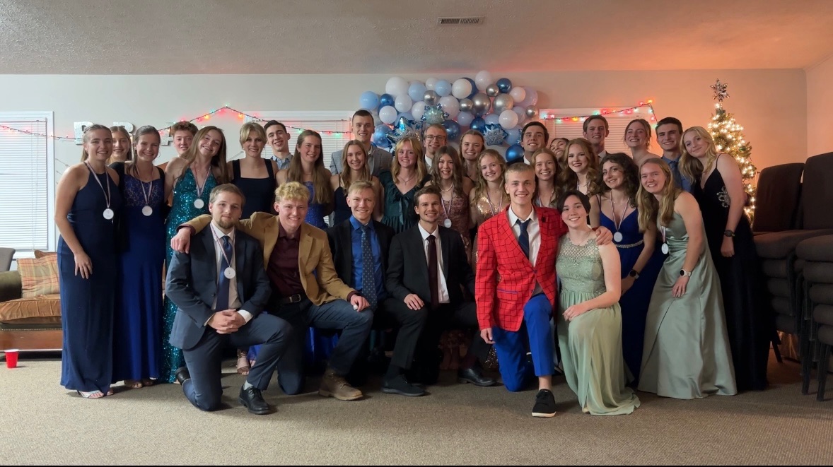 Large group of people in formal attire posing together at an indoor holiday celebration with balloon decorations and a lit Christmas tree.