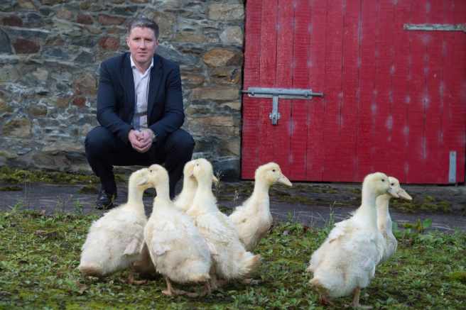 Un homme en costume accroupi devant un groupe de canards blancs près d'une porte rouge en bois.