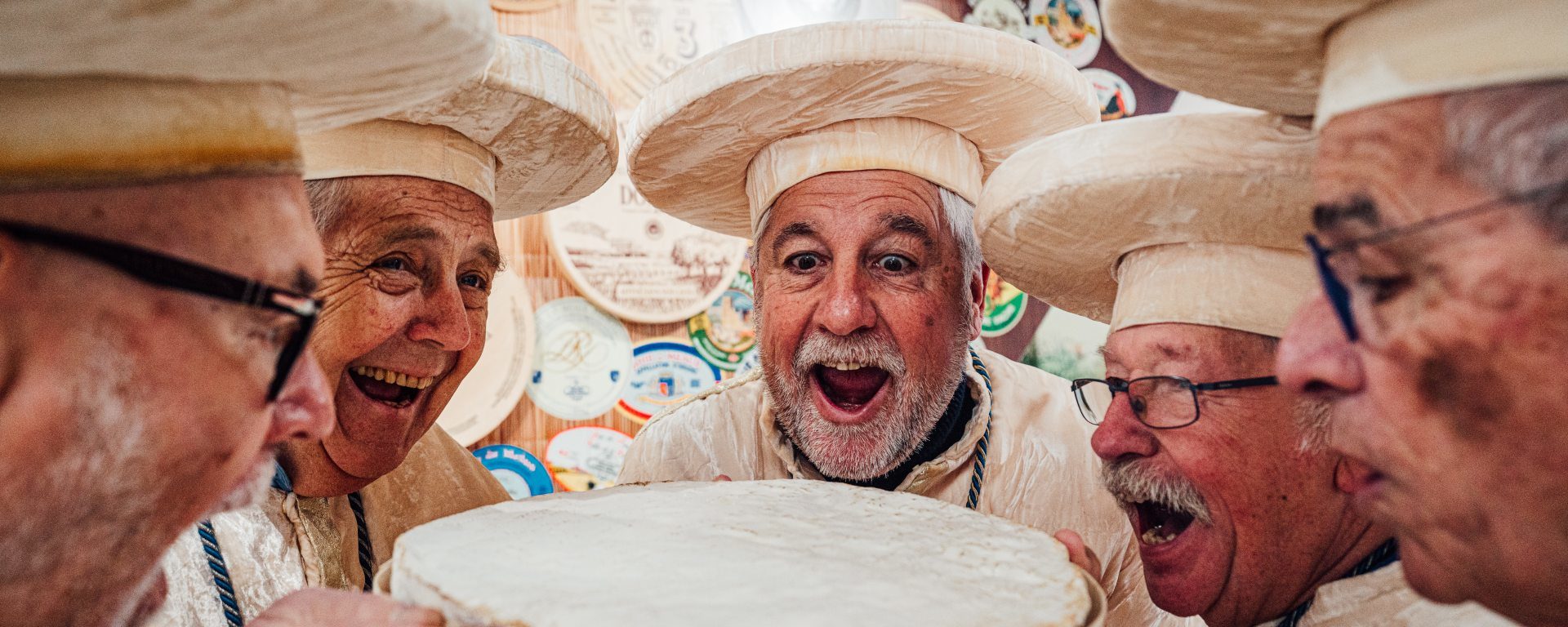 Cinq confrères en tunique et coiffe crème tiennent une meule de brie en riant.