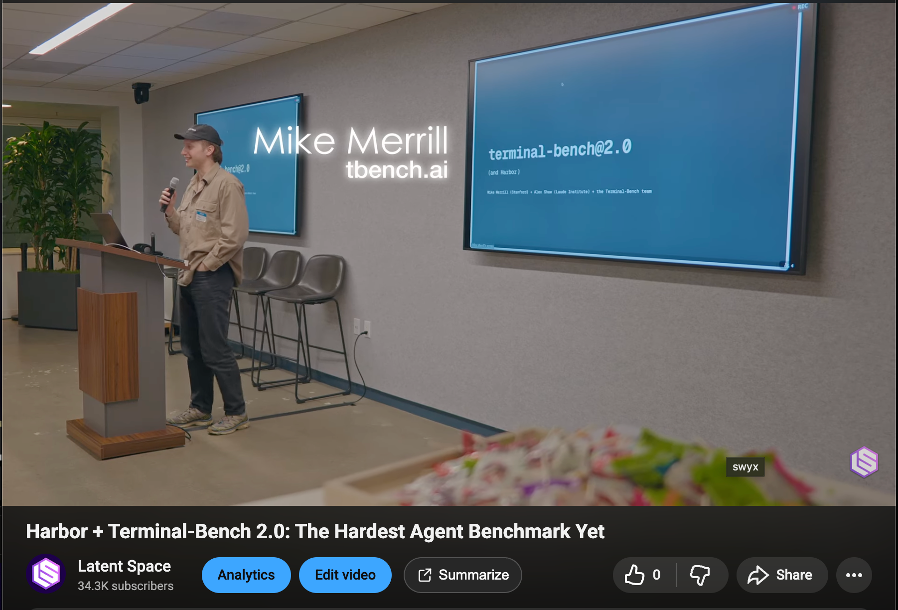 A presenter discusses Terminal-Bench 2.0 at a technical conference, standing at a podium with presentation slides about the AI benchmark.