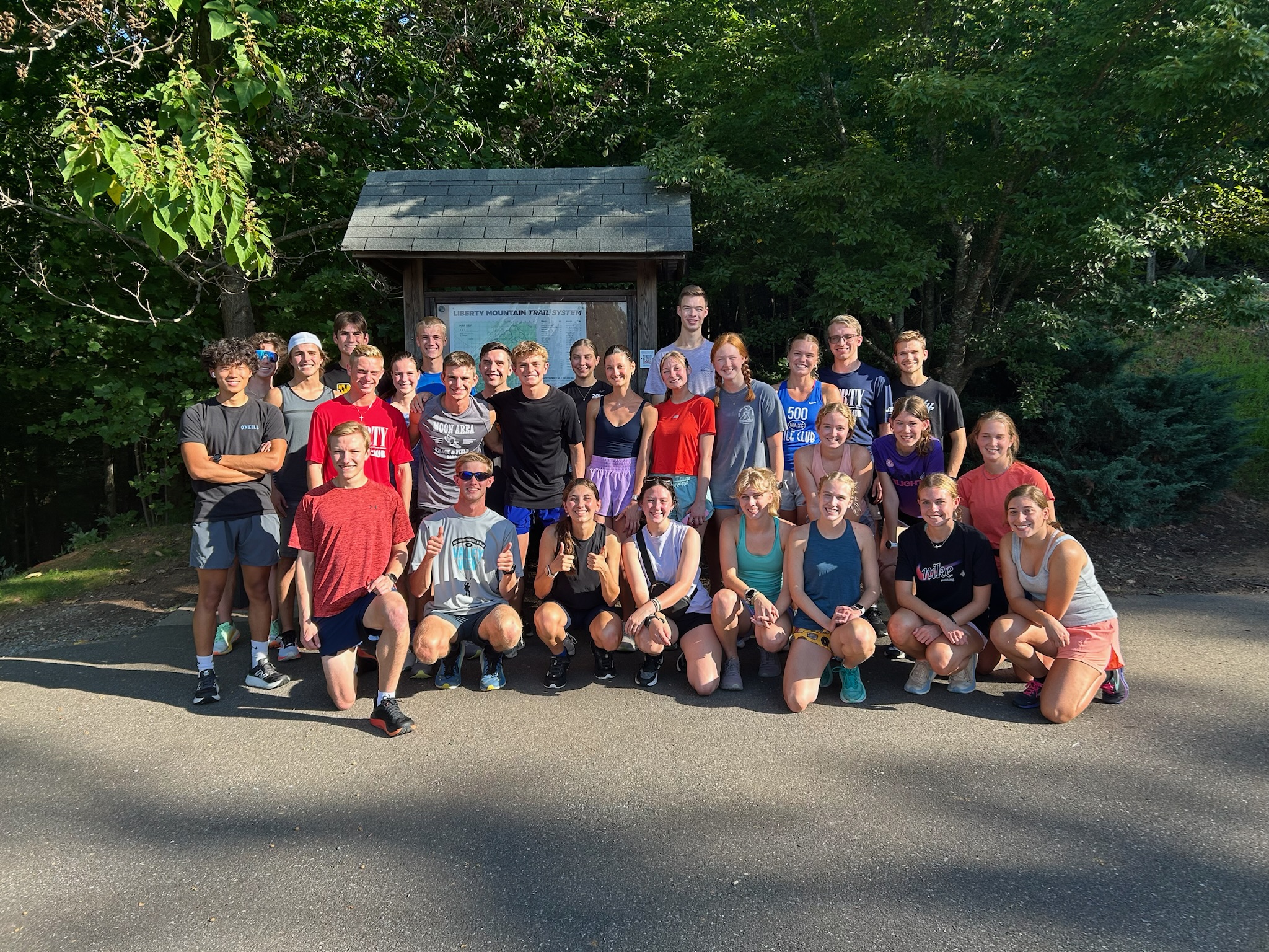 A large group of runners and hikers pose together at a trailhead kiosk surrounded by dense forest.
