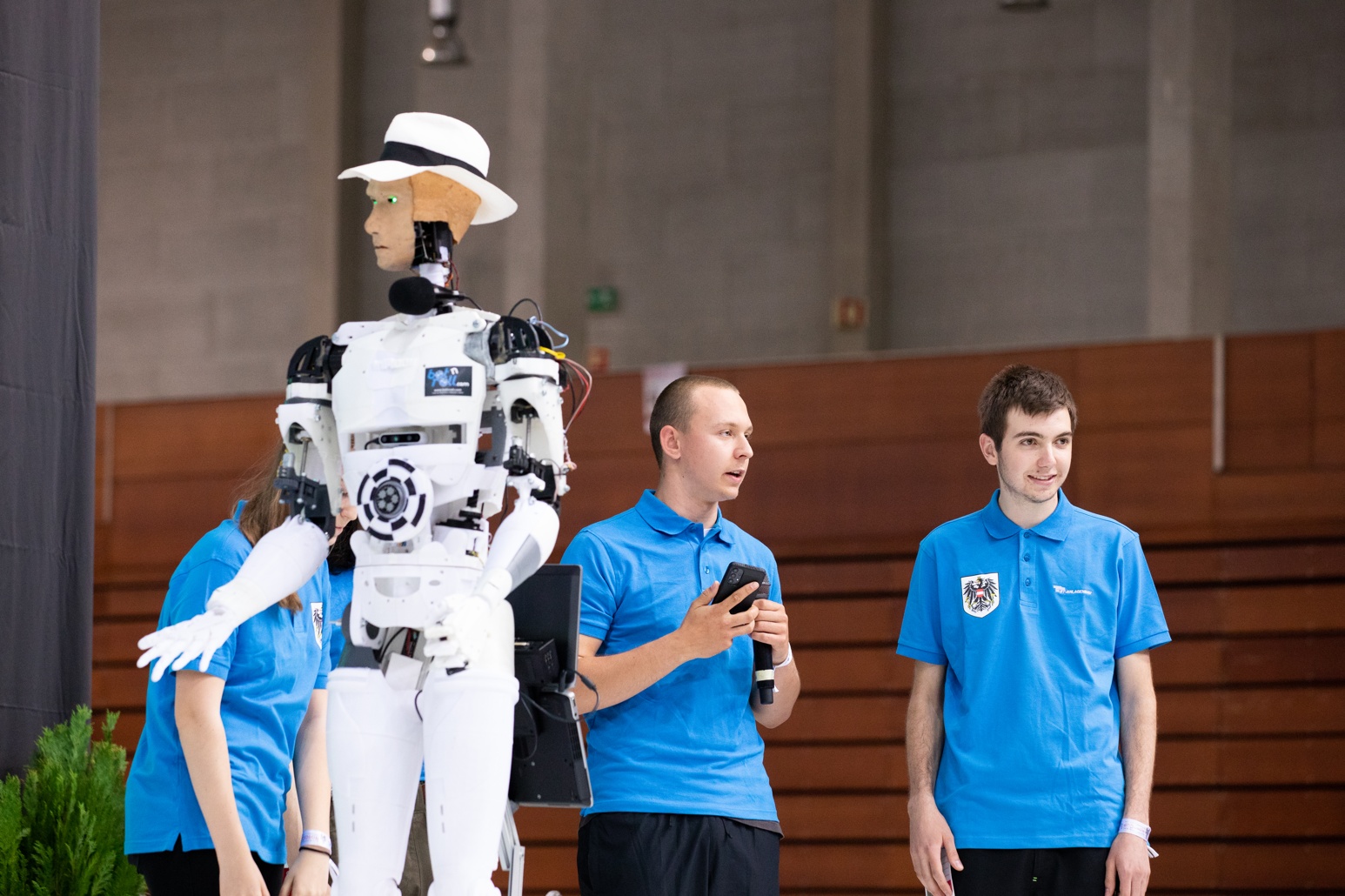 Um robô humanoide com chapéu branco está em pé ao lado de dois homens em camisas azuis, um dos quais segura um telefone e