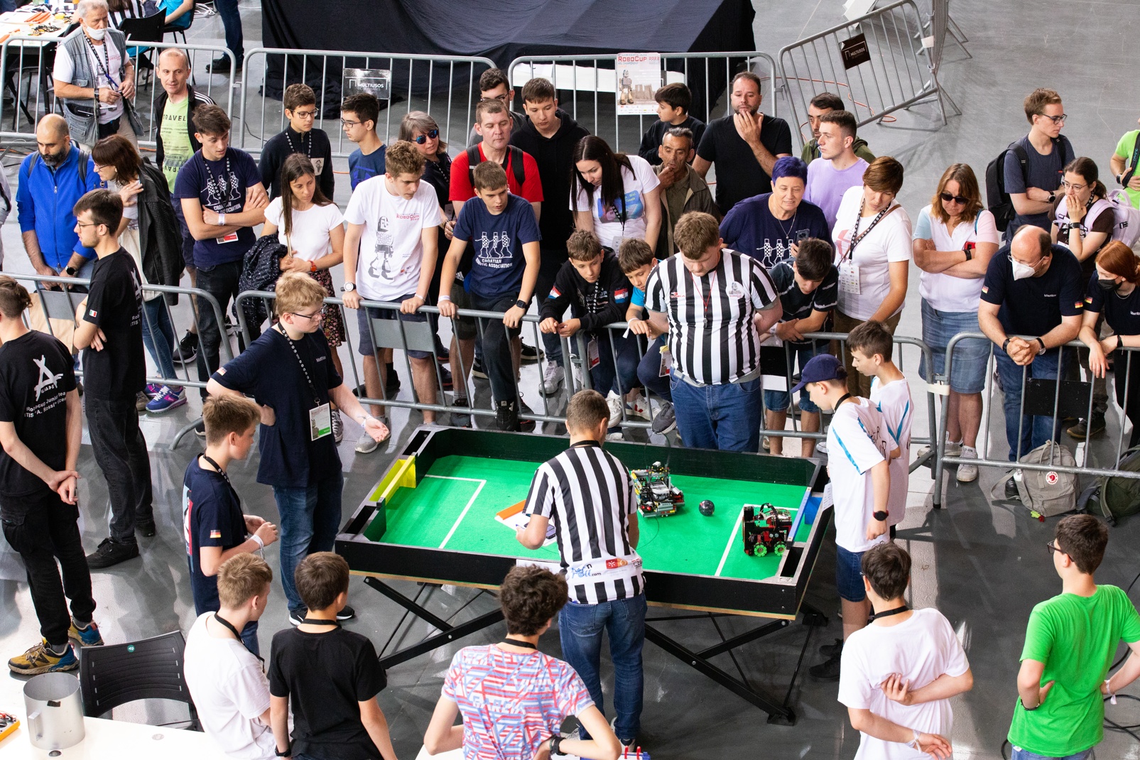 A crowd watches as two small robots compete on a miniature soccer field.