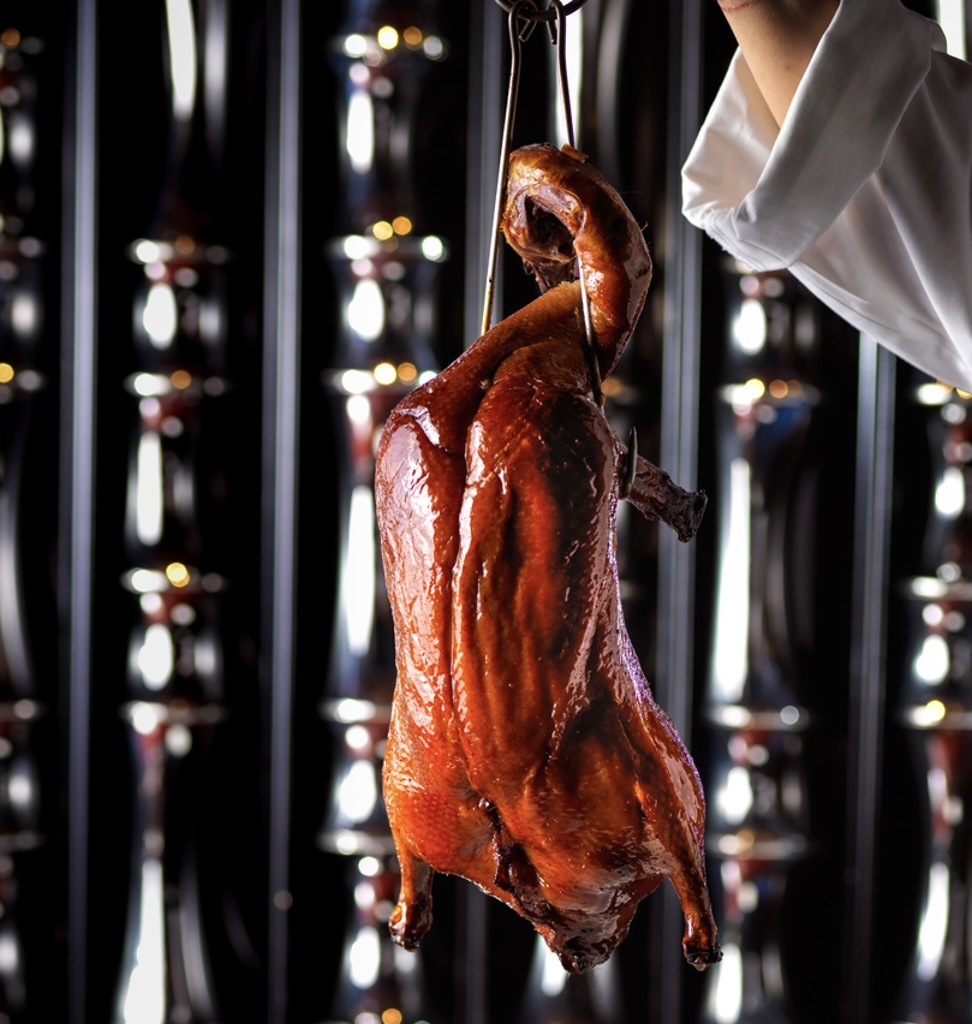 Un chef tient un canard laqué rôti et brillant par le cou, prêt à être découpé.
