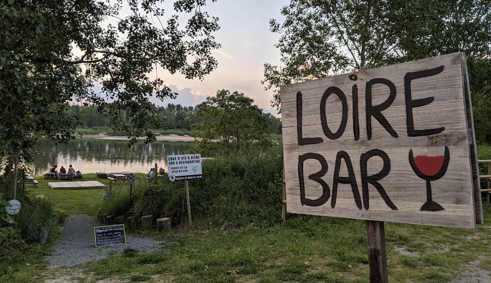 Un panneau en bois indique LOIRE BAR avec un verre de vin rouge peint, au bord de la Loire au crépuscule.