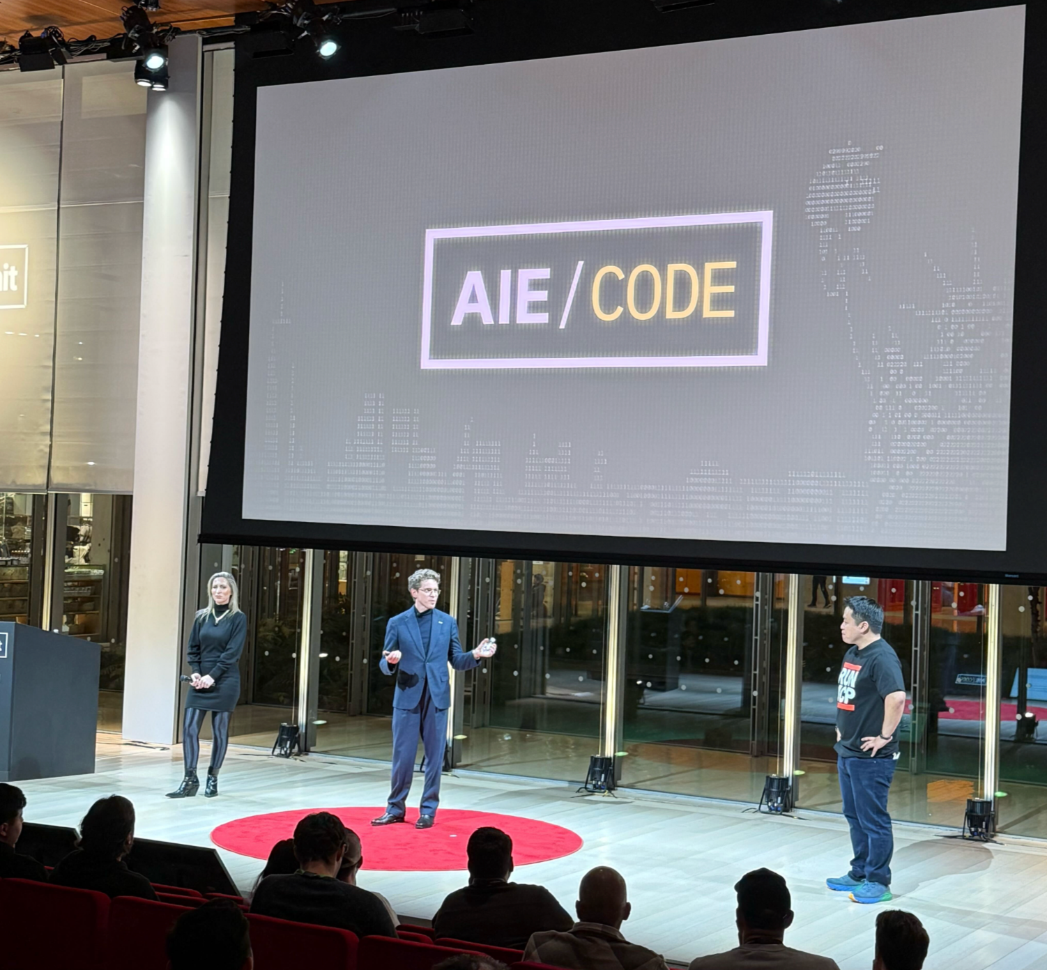 A large screen displaying the "AIE/CODE" logo during a tech conference presentation with speakers on a red circular stage.